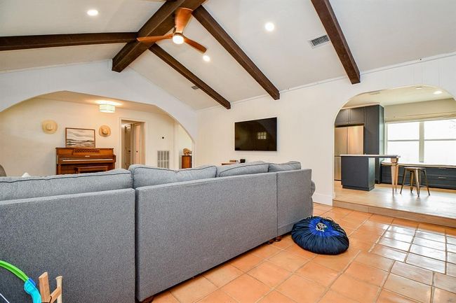 This inviting living space features a cozy sectional sofa, exposed wooden beams, and a ceiling fan. It opens into a kitchen with modern appliances and a breakfast bar. Warm terracotta tiles complement the open layout, creating a comfortable and stylish atmosphere. | Image 15