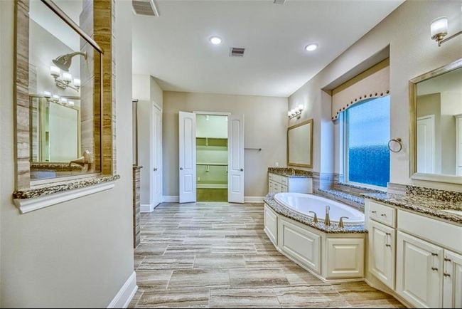 This is a spacious, elegant bathroom featuring a large soaking tub, dual vanities with granite countertops, and ample cabinetry. The room is well-lit with recessed and wall lighting, and has a walk-in shower and closet. The wood-style tile flooring adds a warm touch. | Image 28
