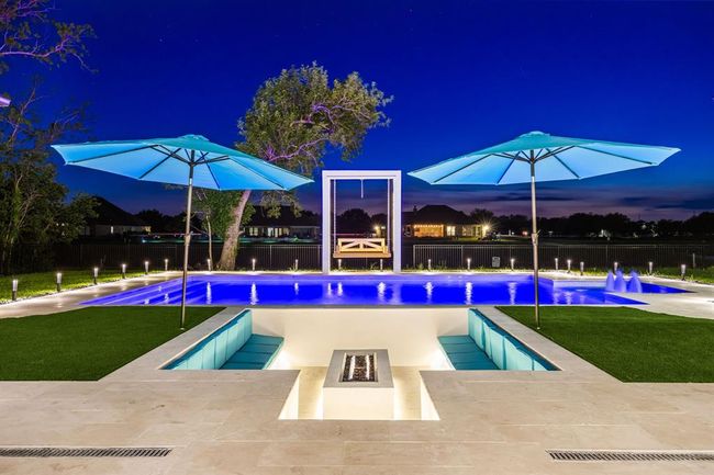 Experience luxury outdoor living with this breathtaking poolscape, featuring a glowing resort-style pool with water features and ambient lighting. A sunken fire pit lounge offers a cozy spot for evening gatherings, while oversized umbrellas provide shade and comfort. The modern swing and expansive backyard views complete this stunning space, perfect for entertaining or unwinding under the stars. | Image 16