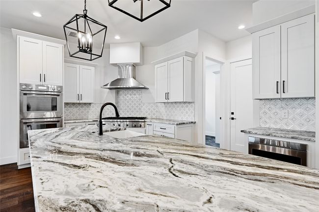 Kitchen featuring dark wood finished floors, white cabinetry, stainless steel double oven, wall chimney exhaust hood, and light stone countertops | Image 10