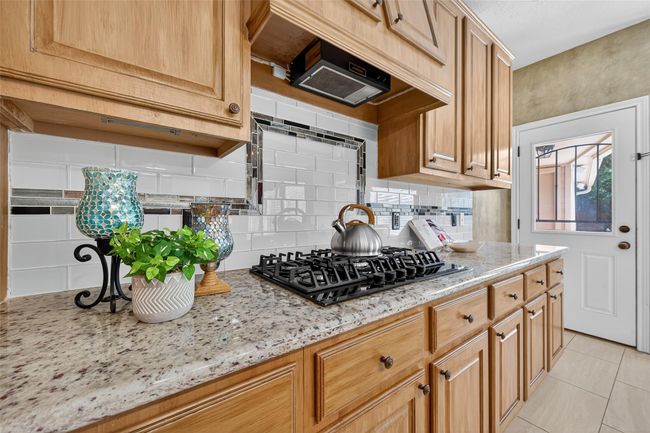 This kitchen features wooden cabinets, a granite countertop, and a sleek gas stove. The white tiled backsplash with decorative accents adds a touch of elegance. Natural light streams through a nearby door, enhancing the space. | Image 10
