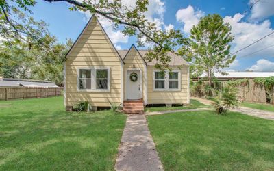 Charming single-story home featuring a peaked roof, light yellow siding, and white trim. The property includes a welcoming front door with a decorative wreath, a well-maintained lawn, and a clear walkway leading up to the house. A wooden privacy fence surrounds the spacious backyard. | Image 1