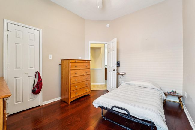 This cozy bedroom features warm wood flooring, neutral walls, and a compact layout with a single bed, wooden dresser, and a small nightstand. It includes a closet and connects to a hallway, providing functional living space. | Image 23