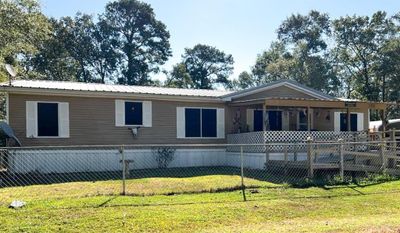 Welcome to your new home, featuring a covered front porch, horizontal siding, and blue window shutters. The property includes a fenced front yard and is surrounded by mature trees, offering a mix of shade and sunny areas. | Image 1