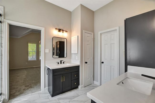 Modern bathroom with sleek black cabinetry, dual sinks, and stylish lighting. Features marble-like flooring and connects to a carpeted room with natural light from a large window. | Image 35