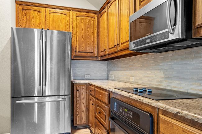Kitchen with black appliances, tasteful backsplash, brown cabinetry, light stone counters, and vaulted ceiling | Image 15