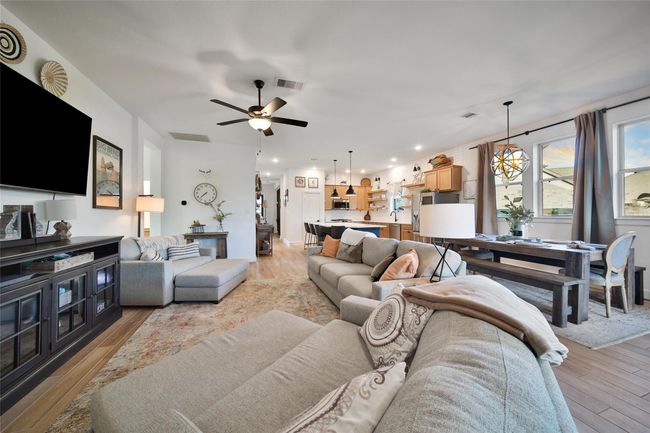 This open-concept living area features a spacious and cozy layout with a large sectional sofa, modern ceiling fan, and stylish decor. The room seamlessly flows into a bright dining area and a well-equipped kitchen with pendant lighting and ample cabinetry. Large windows provide natural light, creating a warm and inviting atmosphere. | Image 27