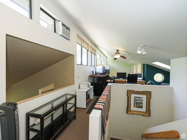 Carpeted living room featuring ceiling fan, vaulted ceiling, an office area, and a wall unit AC | Image 42