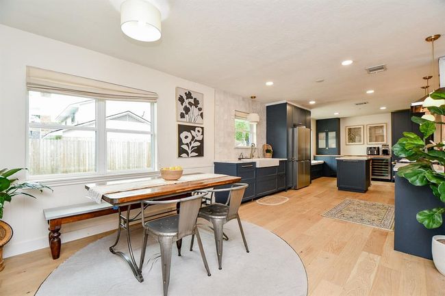 This photo showcases a modern, open-concept kitchen and dining area with light wood flooring. The kitchen features sleek dark cabinetry, stainless steel appliances, and a farmhouse sink. A bright dining space with large windows offers natural light, complemented by a stylish table and chairs. The area is accented with contemporary art and indoor plants, creating a welcoming atmosphere. | Image 24