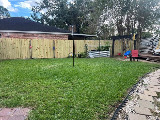 This backyard features a well-maintained lawn, a wooden fence for privacy, and a small deck area. There's a pergola with seating and a garden bed for planting. A playful swing set adds a family-friendly touch. | Image 7