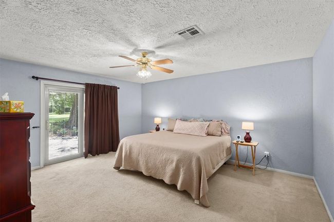 This spacious bedroom features soft carpet, and a ceiling fan. It has a sliding glass door that opens to a serene outdoor view, complemented by light walls and cozy lighting. | Image 20