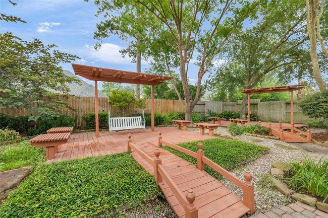 The charming pergola-covered swing sits on a raised wooden platform, surrounded by benches, greenery, and gravel walkways—creating a relaxing backyard oasis. | Image 38
