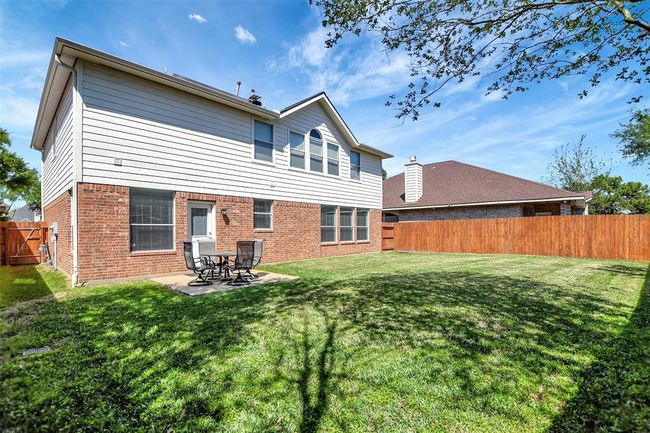 This photo showcases a spacious backyard with a well-maintained lawn and a two-story house featuring brick and siding exterior. There's a small patio area with outdoor seating, ideal for relaxation. The yard is enclosed by a wooden fence, offering privacy and security. | Image 46