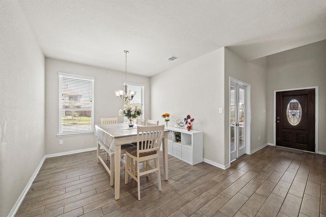 This photo showcases a bright dining area with wood-look tile flooring. It features a table set for six under a tasteful chandelier. Large windows provide ample natural light, and there's a stylish entryway with double glass doors nearby. | Image 8