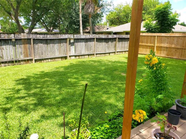 Spacious backyard with lush green grass, bordered by a wooden fence. Features a small patio with potted plants and vibrant yellow flowers, perfect for outdoor relaxation. | Image 13
