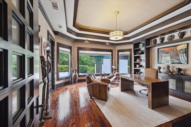 This richly appointed study features custom millwork, patio door, tray ceilings with layered wood molding, and hand scraped wood flooring that adds timeless elegance. A wall of windows frames peaceful pool and garden views, filling the space with natural light and creating an inspiring environment for productivity or relaxation. Built-in shelving and display cabinetry offer both style and function, which also provides a perfect setting for using this space as a first-floor game room. | Image 21