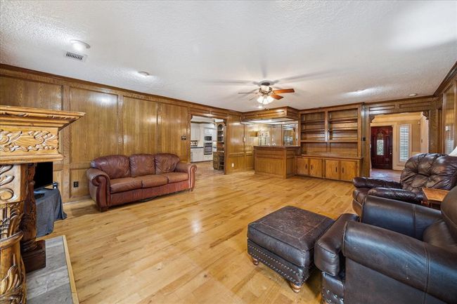 Spacious living room with warm wood paneling, built-in shelving, and a cozy fireplace. Features hardwood flooring, ceiling fan, and ample natural light. Adjacent to the kitchen, ideal for entertaining. | Image 8
