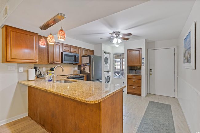 Kitchen featuring brown cabinetry, stacked washer and dryer, a peninsula, light stone counters, and appliances with stainless steel finishes | Image 4