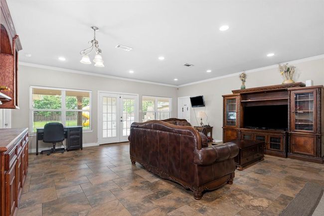 This is a spacious living area featuring tile flooring, ample natural light from large windows and glass doors leading to the outside, a ceiling fan with lighting, and a traditional entertainment center with a large TV space. The room is furnished with a leather sofa and includes a workspace with a desk and chair. | Image 16