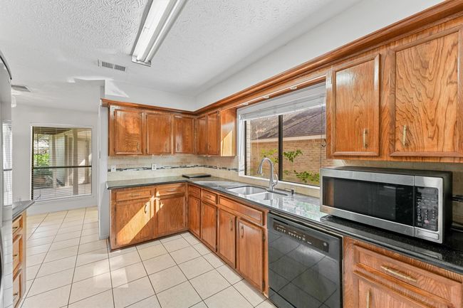 This kitchen features warm wooden cabinetry, a large window over the sink providing natural light, black appliances including a dishwasher and microwave, and light tile flooring. It's bright and inviting, perfect for any home buyer. | Image 4