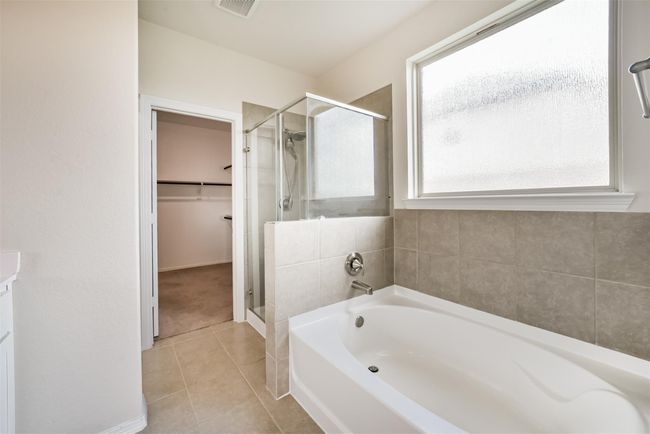 This primary bathroom features a modern design with a spacious bathtub and a separate glass-enclosed shower. It has neutral tile flooring and a large frosted window for natural light. Adjacent is a walk-in closet for convenient storage. | Image 12