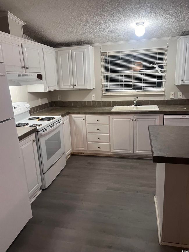 Kitchen featuring white appliances, a textured ceiling, under cabinet range hood, white cabinets, and dark countertops | Image 12
