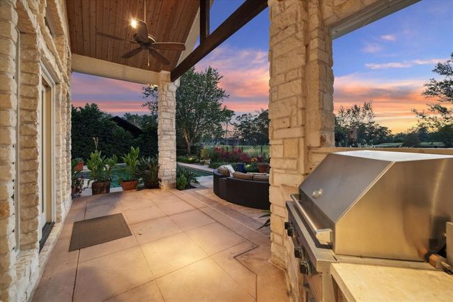 This photo showcases a beautiful outdoor patio with stone walls, a ceiling fan, and a built-in grill. It opens to a spacious garden area with lush greenery and a stunning sunset view, perfect for entertaining or relaxing. | Image 32