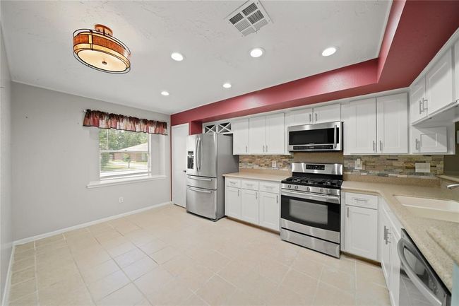 This modern kitchen features stainless steel appliances, white cabinetry, and a tile floor. A window provides natural light, complemented by recessed lighting and a stylish ceiling fixture. The room is accented with red trim and a stone backsplash. | Image 16
