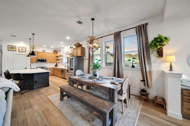 This photo showcases a modern, open-concept kitchen and dining area. It features light wood flooring, a large island with bar seating, and stainless steel appliances. The dining space includes a rustic wooden table, large windows with natural light, and stylish lighting fixtures, creating a warm, inviting atmosphere. | Image 23