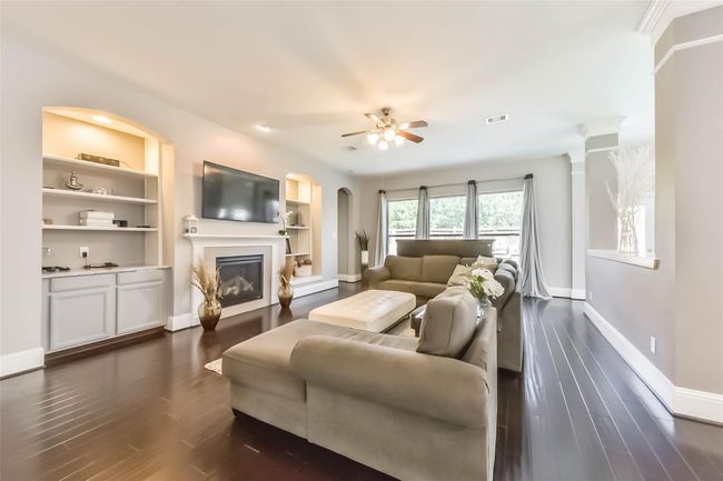 The spacious living room features elegant hardwood floors, a cozy fireplace with built-in shelves, and ample natural light from large windows. The neutral color palette and modern ceiling fan create a welcoming atmosphere. | Image 8