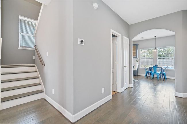 This photo showcases a bright, modern interior with hardwood floors and neutral walls. It features a staircase, a hallway leading to a kitchen area with large windows, and a cozy dining space with blue chairs. | Image 25