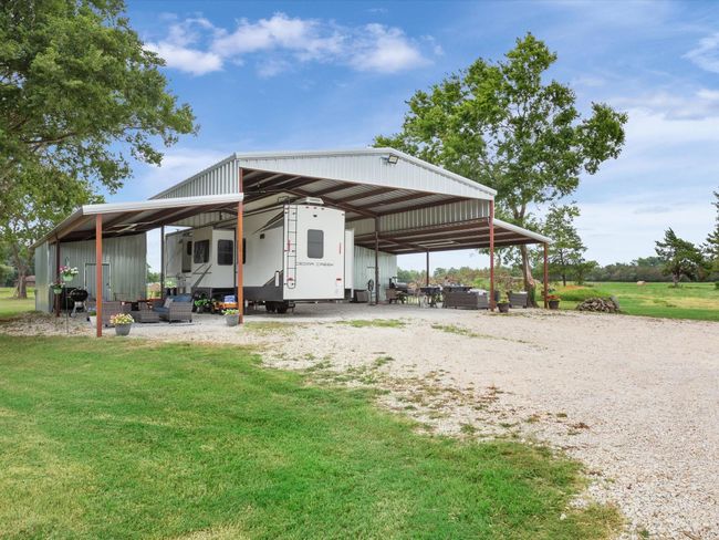 A recreational vehicle parked under a large metal carport surrounded by open green space and trees on a clear day. | Image 10