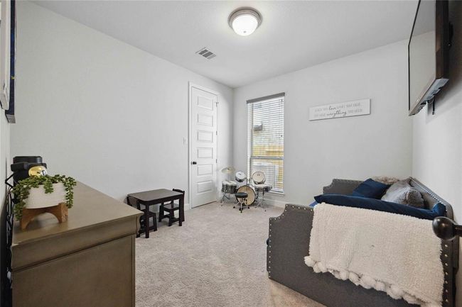 Cozy first floor bedroom with neutral walls, carpet flooring, and a ceiling light. Features a small drum set by the window, a cushioned bench with pillows, and a dresser with decorative plants. Ideal as a child's playroom or small bedroom. | Image 19