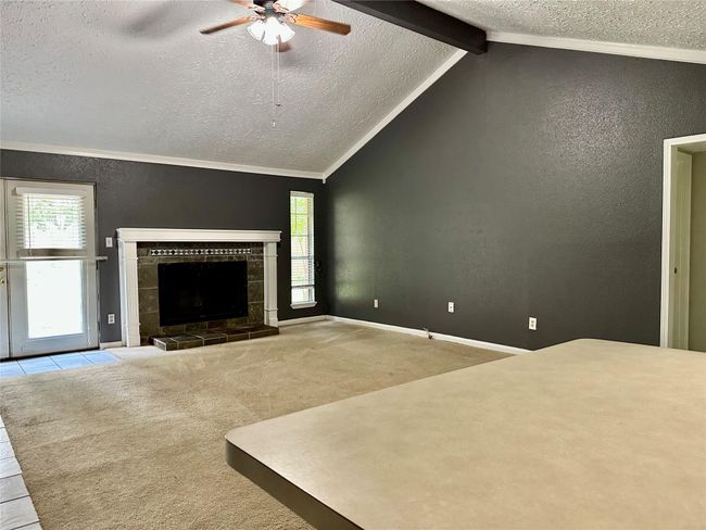 Spacious living room with vaulted ceilings and a ceiling fan, featuring a cozy fireplace. The room has neutral carpet flooring and a dark accent wall, complemented by natural light from the windows and a glass door leading outside. | Image 4