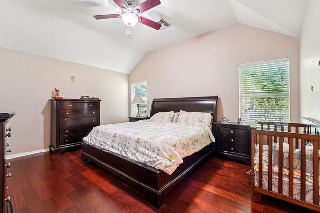 This spacious bedroom features hardwood floors, a vaulted ceiling with a fan, and ample natural light from two windows. It includes a large bed, dark wood furniture, and a crib, creating a cozy and functional space. | Image 14