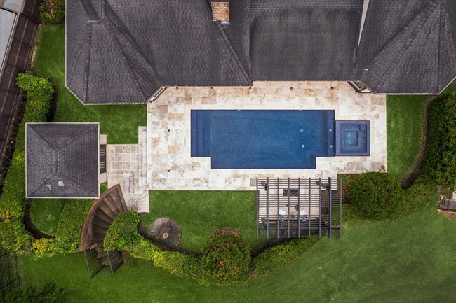 This aerial perspective reveals the meticulously planned backyard oasis, where symmetry and design blend seamlessly. At the heart lies a sleek, rectangular saltwater pool with an integrated spa, bordered by creamy travertine decking that enhances the clean lines of the space. A contemporary pergola with a cozy fire pit anchors one end, while the pool house and gracefully curved staircase to the creek and greenspace beyond create a sense of harmony and balance within the manicured landscape. | Image 48