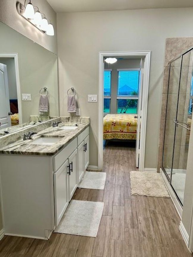 This bathroom features a dual-sink vanity with a stylish granite countertop, a spacious glass-enclosed shower, and tile flooring. It connects to a bedroom with large windows, providing ample natural light. | Image 15