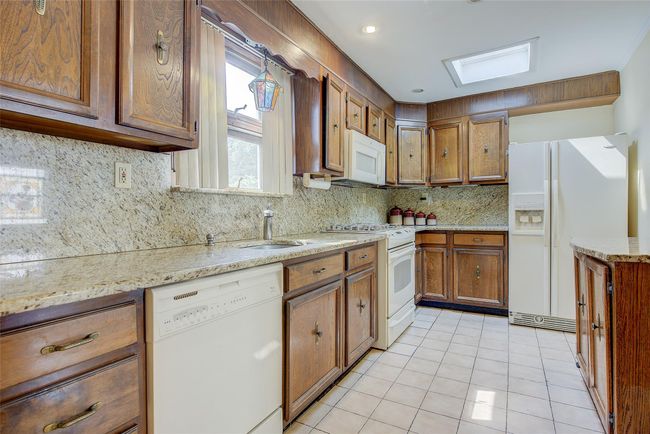 Kitchen featuring white appliances, brown cabinets, light tile patterned floors, tasteful backsplash, and light stone counters | Image 13