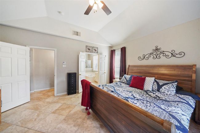 This spacious bedroom features a vaulted ceiling and tiled flooring. It includes an ensuite bathroom with double doors, and natural light filters through two windows with red curtains, creating a cozy and inviting atmosphere. | Image 19