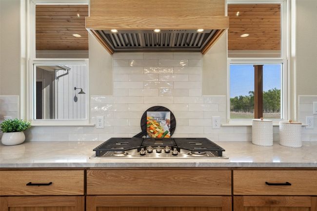 Kitchen featuring custom exhaust hood, stainless steel gas stovetop, decorative backsplash, light stone countertops, and brown cabinetry | Image 12