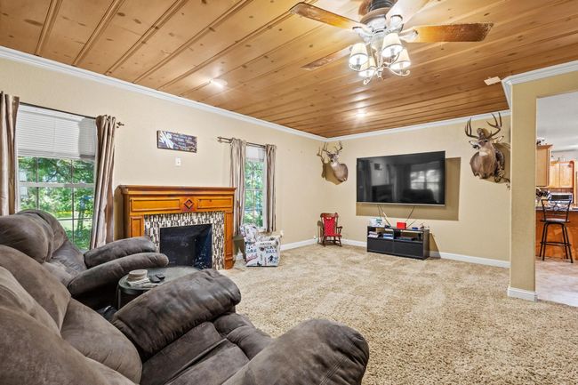 Carpeted living area featuring a tiled fireplace, baseboards, ceiling fan, crown molding, and wooden ceiling | Image 6