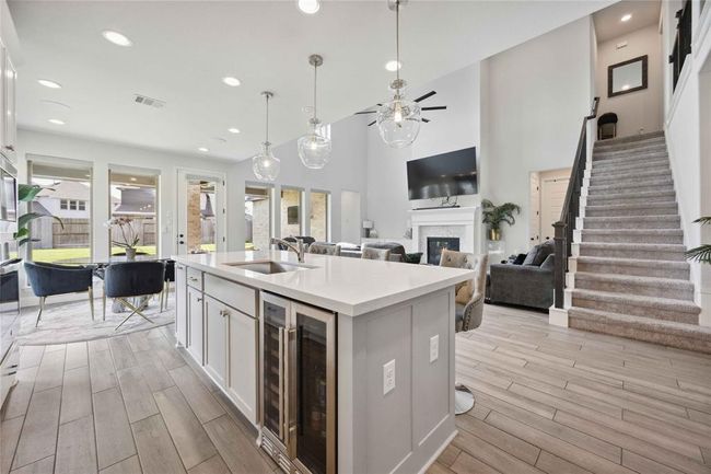 This spacious and modern open-concept living area features a bright kitchen with an island, stylish pendant lighting, and large windows offering plenty of natural light. Adjacent is a cozy living room with a fireplace and media setup, complemented by a sleek staircase leading to the upper level. | Image 18