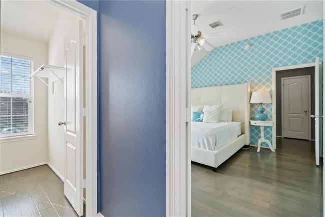 This photo shows a bright, stylish bedroom with a modern blue accent wall and hardwood flooring. It features a cozy bed with decorative pillows and a small bedside table with a lamp. On the left, there's a walk-in closet with a window, providing natural light. | Image 31