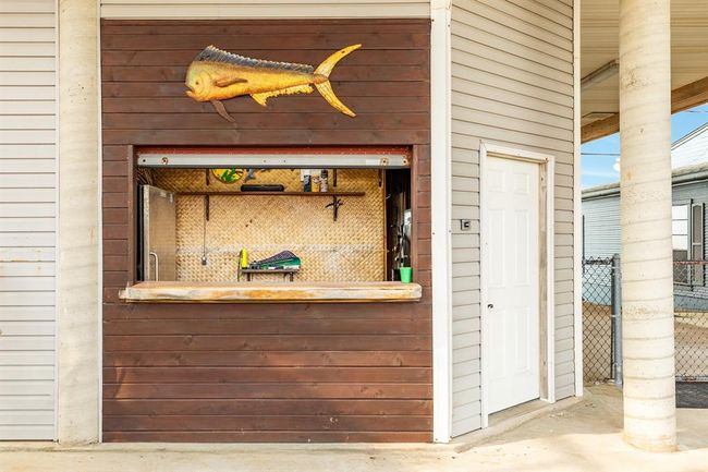 This photo showcases an outdoor bar area with a wooden facade, featuring a fish decoration. It includes a serving counter with a backsplash and a nearby door leading into the garage ideal for entertaining. | Image 42