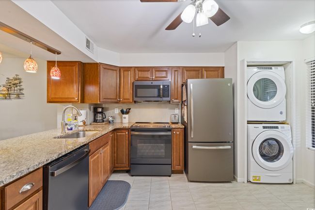 Kitchen with brown cabinetry, stacked washer and clothes dryer, light stone countertops, a sink, and appliances with stainless steel finishes | Image 8