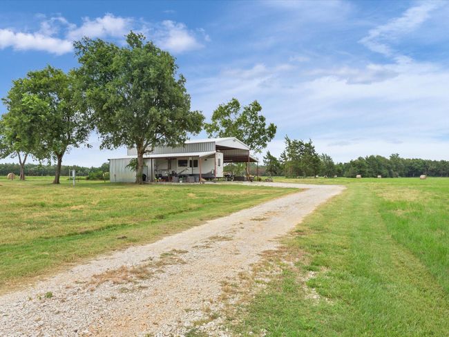 Rural landscape with a gravel driveway leading to a custom metal RV barn, flanked by trees and a field with hay bales under a cloudy sky. | Image 6