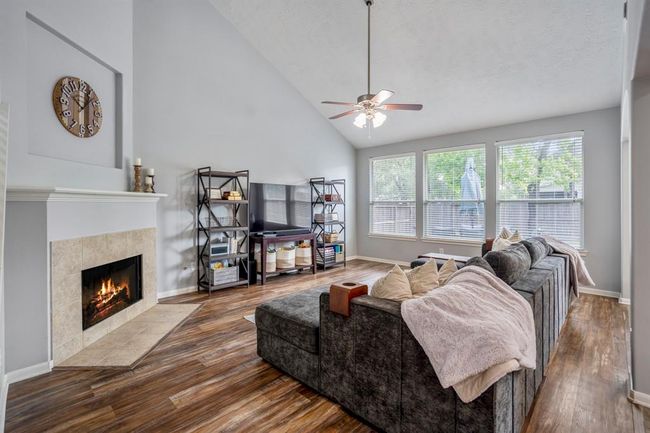 The spacious living room boasts a two-story vaulted ceiling, creating an open and airy feel. A corner fireplace with a trimmed mantle and a recessed art niche above add a touch of elegance. Large windows offer a serene view of the backyard, flooding the space with natural light. | Image 6
