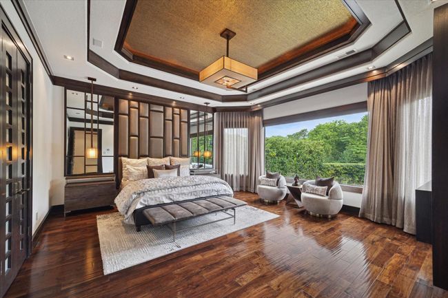 This opulent primary bedroom exudes calm and sophistication, featuring a custom upholstered wall flanked by wood-trimmed mirrors, high-end designer lighting, and a dramatic tray ceiling with inlaid gold leaf accents. A wall of windows covered with lavish draperies frame lush, tree-lined views of the backyard oasis while flooding the space with natural light. The intimate sitting area is ideal for morning coffee or evening unwinding, making this suite the ultimate retreat for rest and relaxation. | Image 25