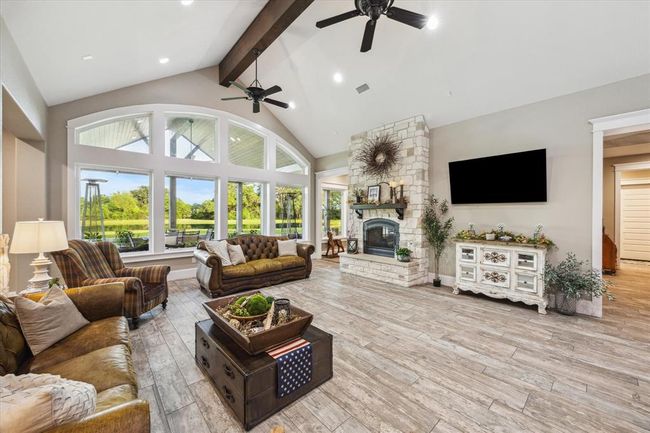 The expansive living room features a stunning double-sided gas fireplace, solid wood beams accentuating the 12-foot ceilings, and custom floor tiles throughout. A dramatic wall of windows offers sweeping views of the covered patio and serene pond, seamlessly blending indoor and outdoor living spaces. | Image 4