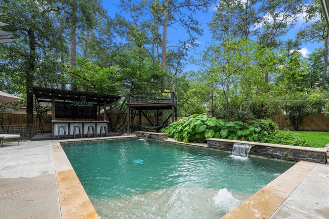 This backyard oasis features a stunning pool with a waterfall, surrounded by lush greenery and a wooden pergola. There's a poolside bar area with an area for stool seating, perfect for entertaining, set against a backdrop of tall, mature trees for added privacy. | Image 36